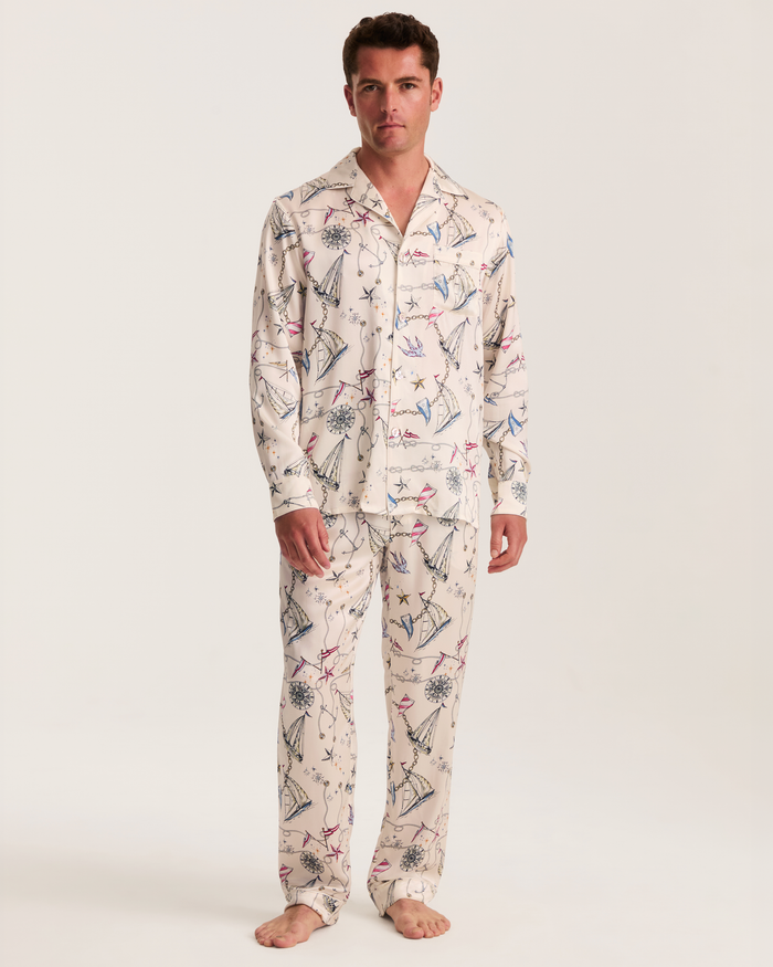 A man stands barefoot wearing the Men’s Silk Pajama Set in Regatta Royale, featuring matching cream-colored pajamas adorned with a colorful, playful print of umbrellas and flowers on a plain white background.