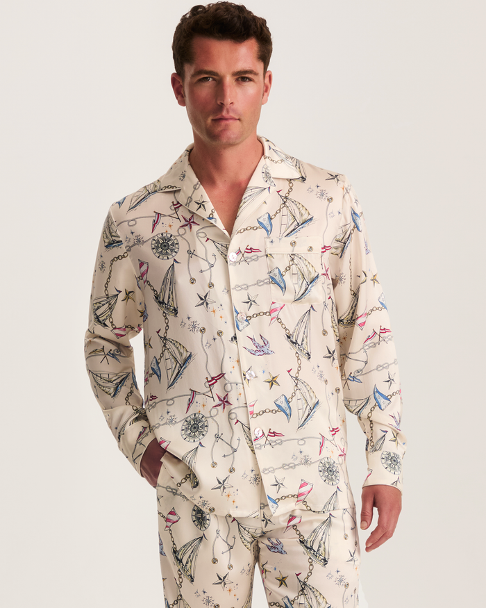 A man models the Men’s Silk Pajama Set in Regatta Royale, featuring a cream base adorned with a sailing-inspired print of boats, compasses, and ropes. He stands against a plain white background, looking at the camera with a neutral expression.
