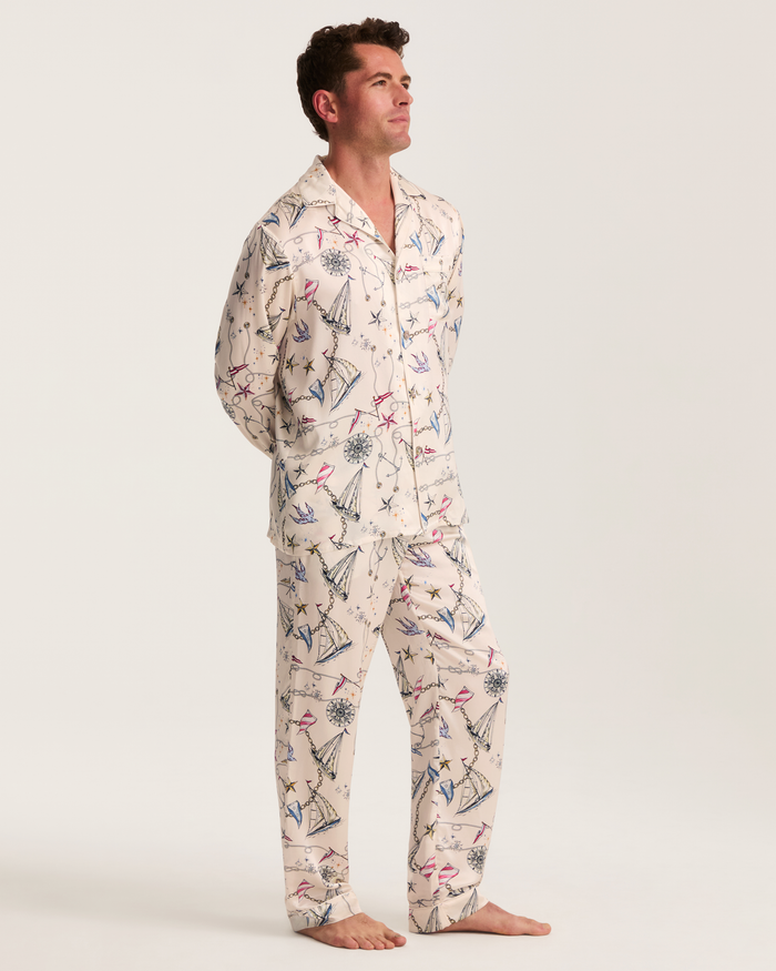 A man stands barefoot against a plain background, dressed in the Men’s Silk Pajama Set in Regatta Royale—cream-colored pajamas featuring a whimsical print of sailboats, anchors, and other nautical motifs—as he gazes slightly upward.