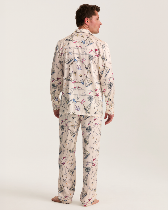 A person stands barefoot and faces away, wearing the Men’s Silk Pajama Set in Regatta Royale—light-colored pajamas adorned with a whimsical pattern of airplanes, stars, and clouds—against a plain white background.