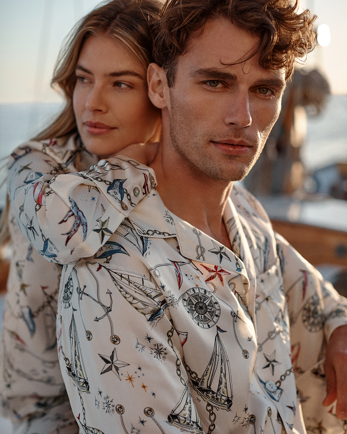 A woman embraces a man from behind on a boat at sunset. Both are wearing matching Men’s Silk Pajama Sets in Regatta Royale, featuring nautical motifs of ships, stars, and compasses. The atmosphere is relaxed and intimate.