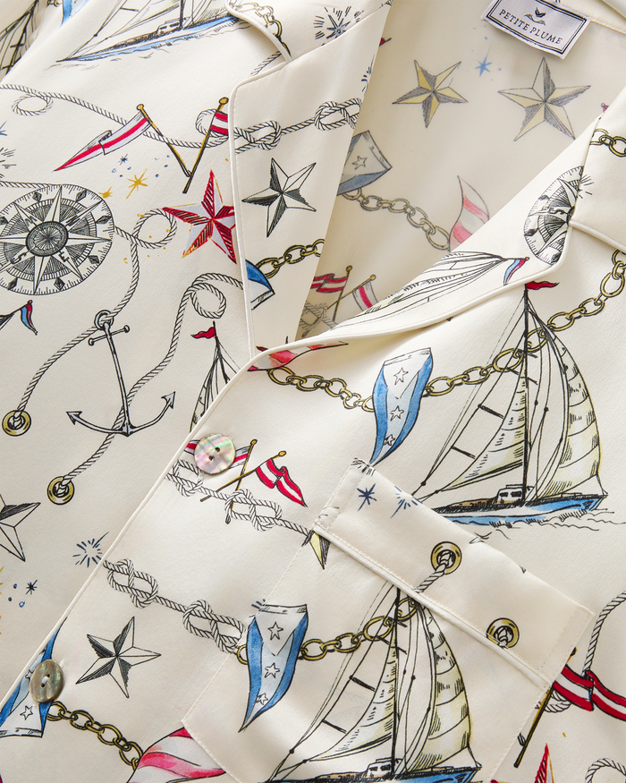 Close-up of the Men’s Silk Pajama Set in Regatta Royale, showcasing a cream-colored fabric adorned with a nautical print of sailboats, anchors, compasses, chains, and stars in shades of blue, red, black, and gold. A button and pocket are visible on the shirt.