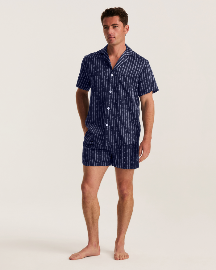A man stands barefoot against a light background, wearing the Men’s Silk Pajama Short Set in Nautical Knots—a matching short-sleeve, button-up navy set with white vertical stripes and coordinating shorts.