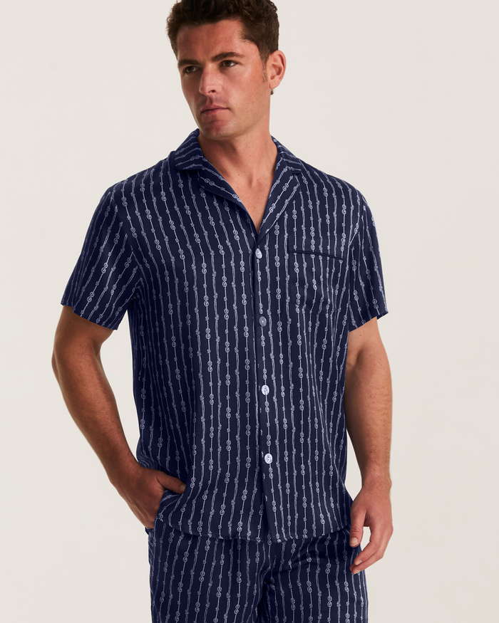 A man wearing the Men’s Silk Pajama Short Set in Nautical Knots—featuring a short-sleeve, button-up, navy blue shirt with white vertical knot patterns and matching shorts—stands with one hand in his pocket against a plain background.