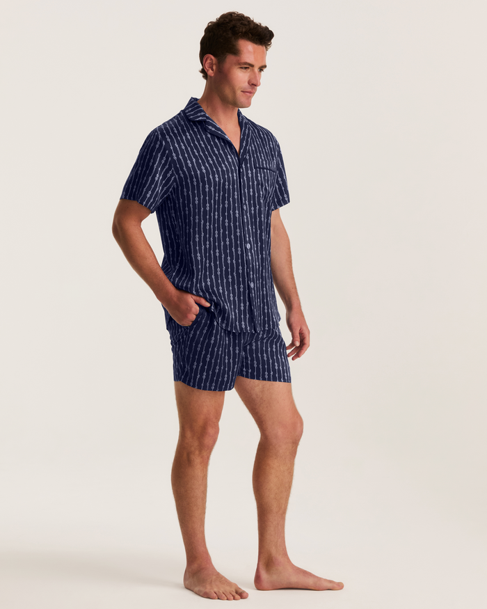 A man stands barefoot against a plain light background, wearing the Men’s Silk Pajama Short Set in Nautical Knots, featuring a matching short-sleeve, button-up navy top and shorts with vertical white stripes.