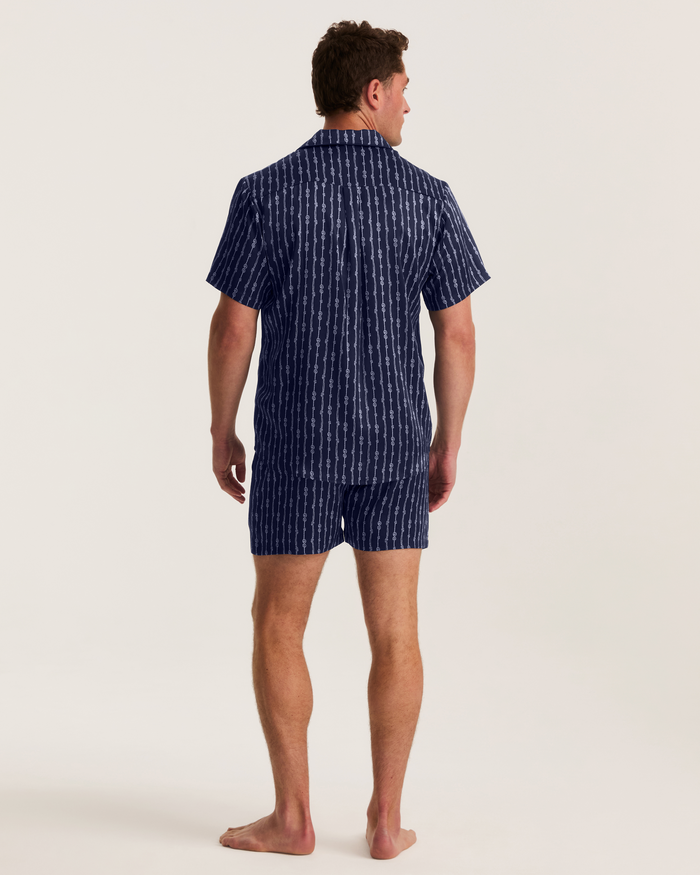 A man stands barefoot facing away, wearing the Men’s Silk Pajama Short Set in Nautical Knots—a matching short-sleeve shirt and shorts set featuring a dark blue and white vertical stripe pattern—against a plain light background.