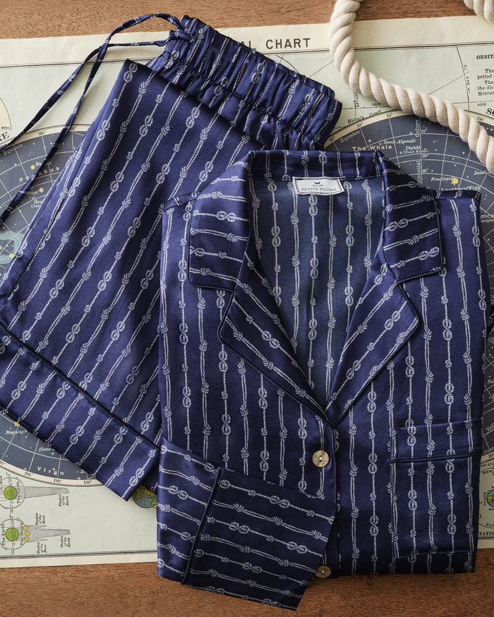 The Men’s Silk Pajama Short Set in Nautical Knots, featuring a navy blue fabric adorned with a white rope knot design, is folded neatly atop a nautical map, accompanied by a matching drawstring bag and a coiled white rope placed nearby.