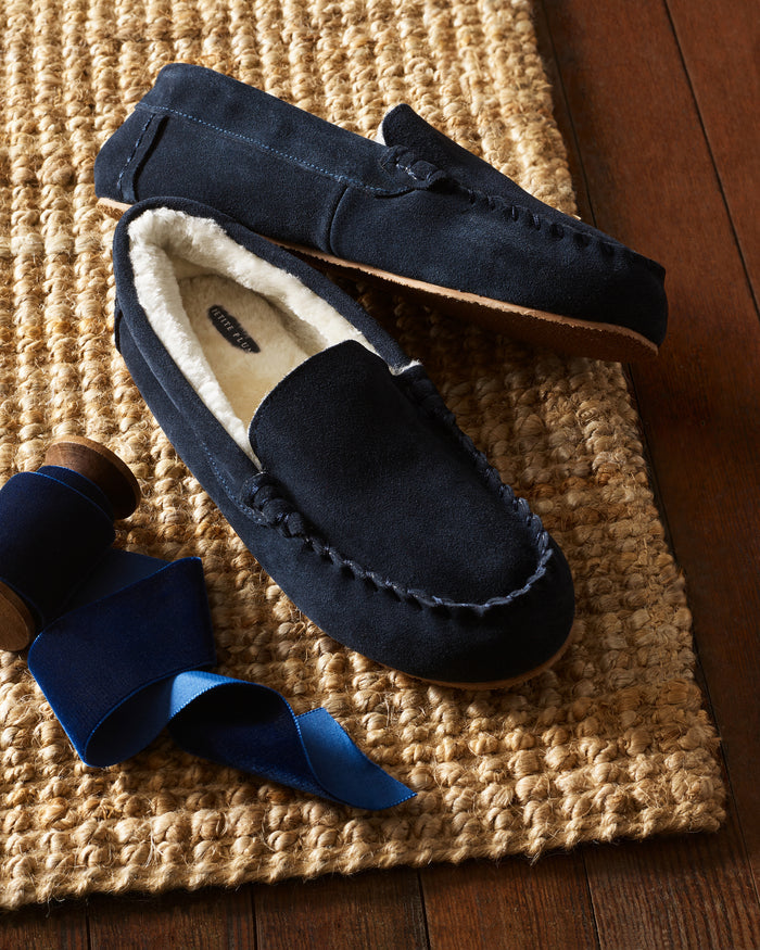 Mens Suede Slippers in Navy by Petite Plume, featuring ultra-soft faux fur lining, rest on a woven mat. Nearby, a spool of dark blue ribbon lies partially unrolled on the rich wooden floor.