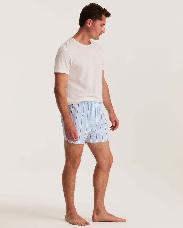 A man stands barefoot on a plain background, wearing a white t-shirt and the Men’s Twill Boxer Shorts in the Coastal Stripe pattern from the 3 Pack set (Coastal Stripe, Anchors Away, and Navy). He is looking to his left with his arms relaxed by his sides.