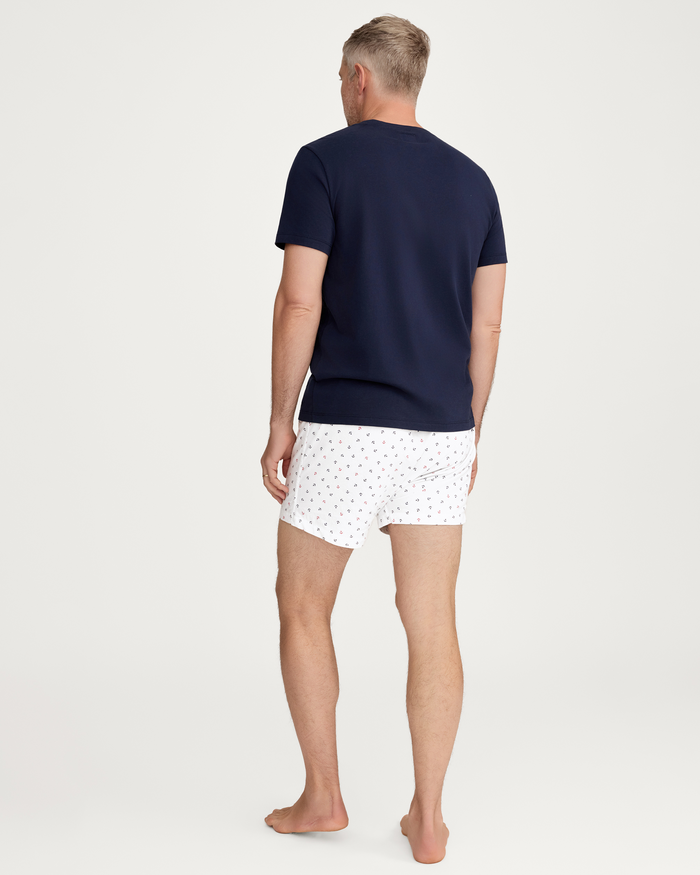 A man stands barefoot on a white background, facing away. He wears a navy blue t-shirt and a pair of men’s twill boxers in the Anchors Away pattern from the Men’s Twill 3 Pack Boxers set, which features Coastal Stripe, Anchors Away, and Navy designs.