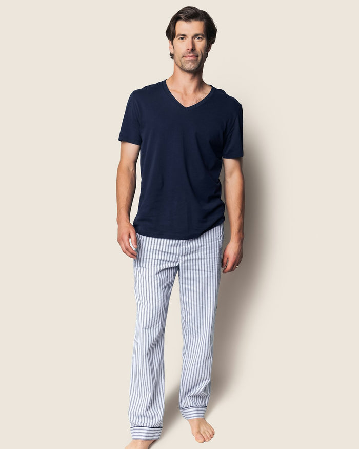 A man stands against a plain background in a navy V-neck T-shirt, paired with the Petite Plume Men’s Twill Classic Navy Pajama Pant Duo in light blue and white stripes. He is barefoot with a neutral expression.