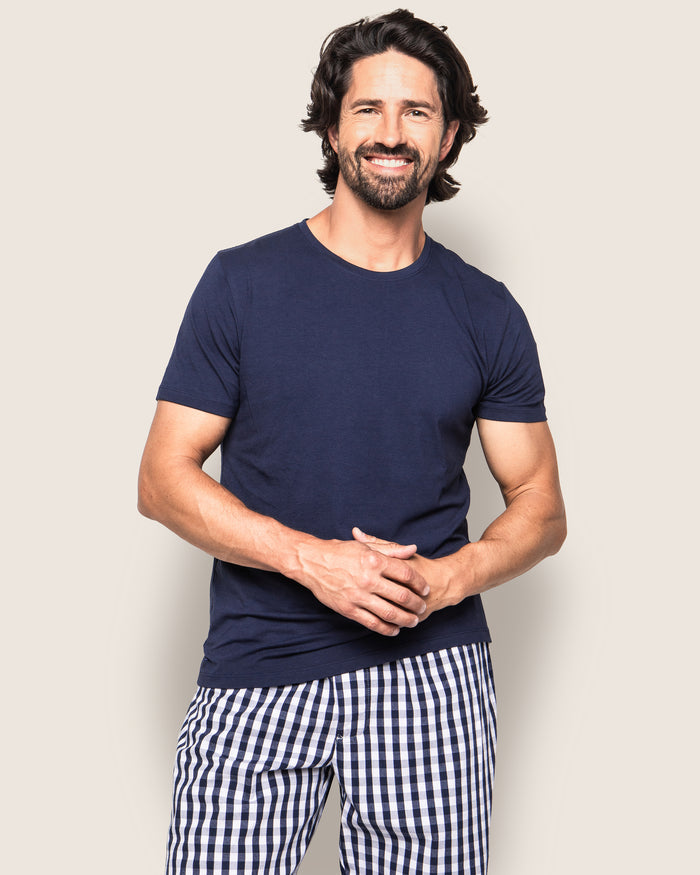 A bearded man with wavy hair is smiling and standing against a light background, wearing a navy blue t-shirt and Mens Twill Pajama Pant in Navy Gingham by Petite Plume, with his hands clasped.