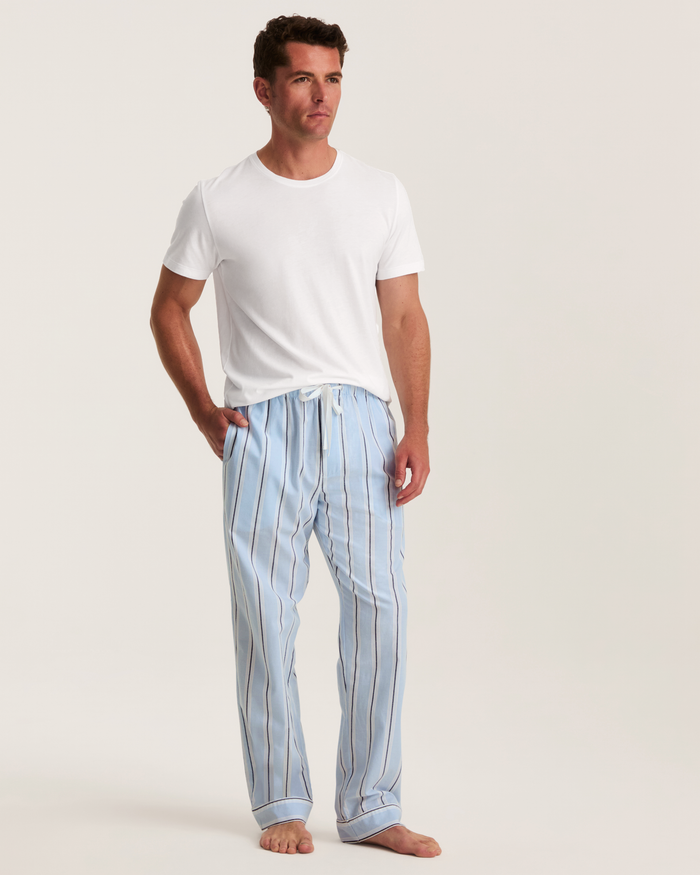 A man stands barefoot wearing a plain white T-shirt and Men’s Twill Pajama Pants in Coastal Stripe, with his hands in his pockets, against a plain white background.