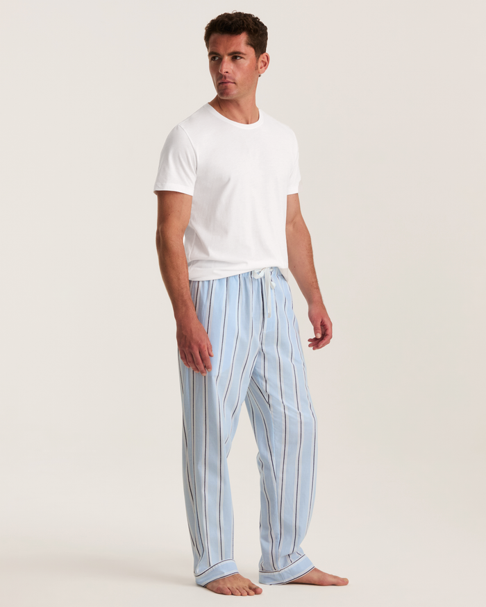 A man stands barefoot against a plain light background, wearing a white T-shirt and the Men’s Twill Pajama Pants in Coastal Stripe, looking off to the side.