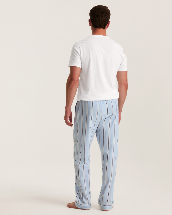 A man stands barefoot facing away, wearing a white T-shirt and Men’s Twill Pajama Pants in Coastal Stripe, against a plain off-white background.