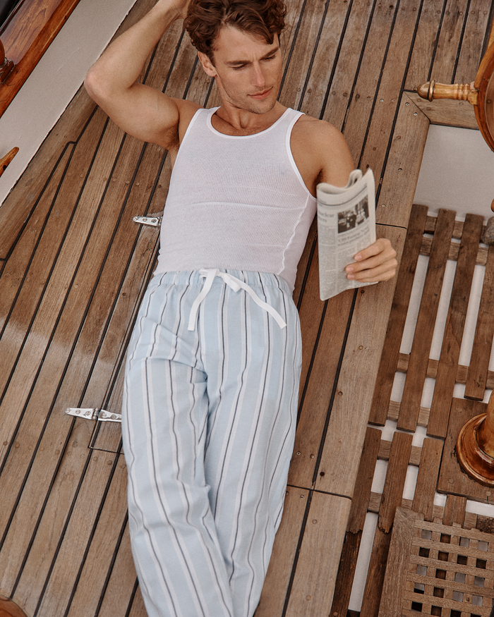 A man in a white tank top and Men’s Twill Pajama Pants in Coastal Stripe lies on a wooden deck, reading a newspaper.