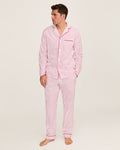 A man stands barefoot wearing the Men’s Twill Pajama Set in Antique Red Ticking, which features matching pink and white stripes with long sleeves and pants. He is looking to his left against a plain, light-colored background.