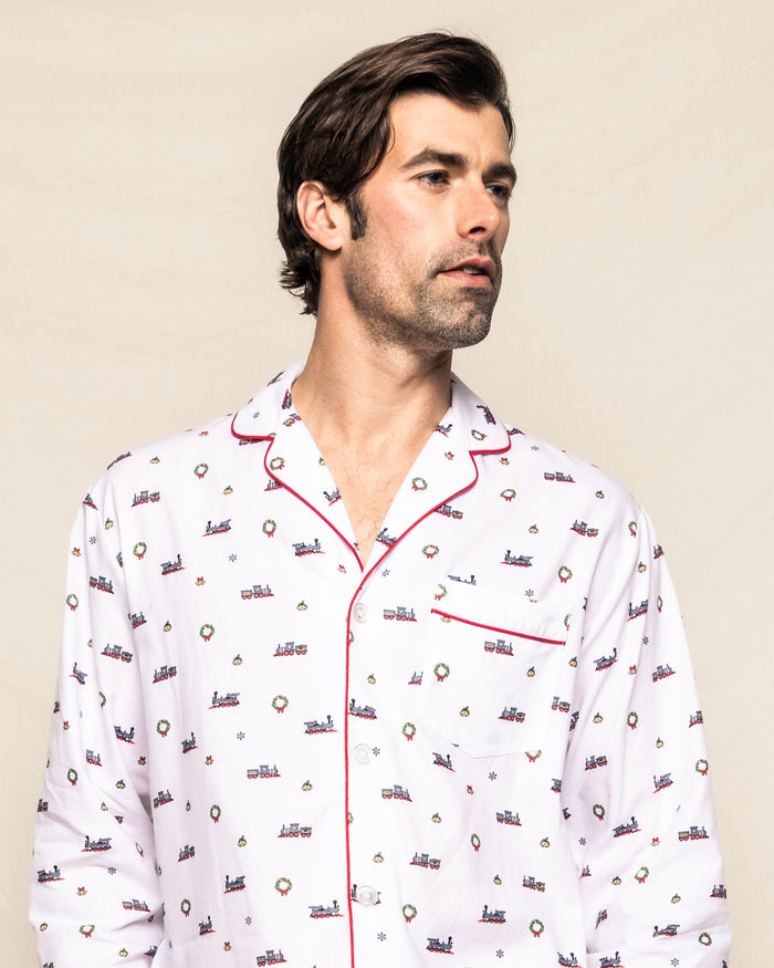 A man in Petite Plumes Mens Twill Pajama Set in Arctic Express gazes sideways against a plain backdrop. The white cotton pajamas, adorned with colorful patterns, boast a red-trimmed collar and pocket, highlighting exquisite yarn-dyed craftsmanship.