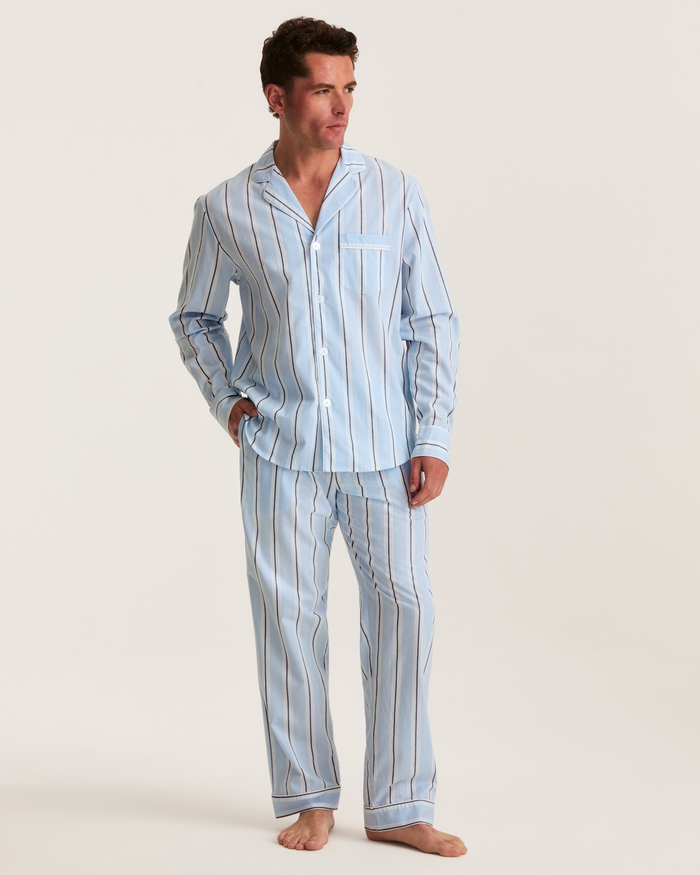 A man stands barefoot on a neutral background wearing the Men’s Twill Pajama Set in Coastal Stripe—light blue, long-sleeved, button-up pajamas featuring vertical white and dark stripes.