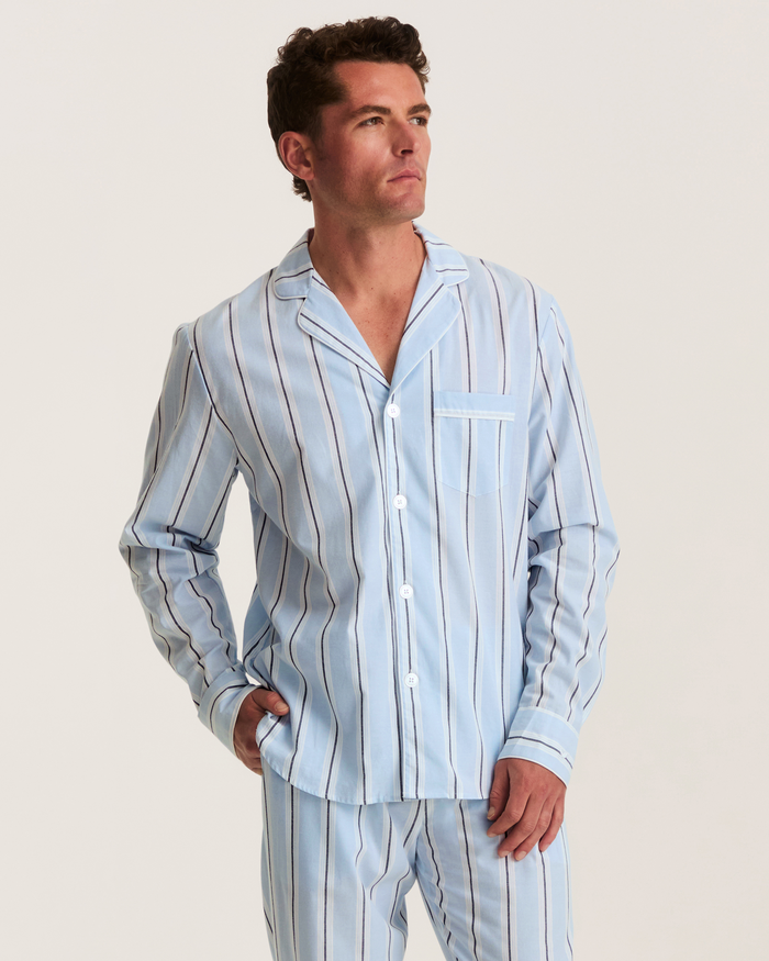 A man models the Men’s Twill Pajama Set in Coastal Stripe—a light blue, long-sleeve, button-up pajama ensemble featuring dark and white vertical stripes—as he stands with one hand in his pocket, gazing off to the side against a plain background.