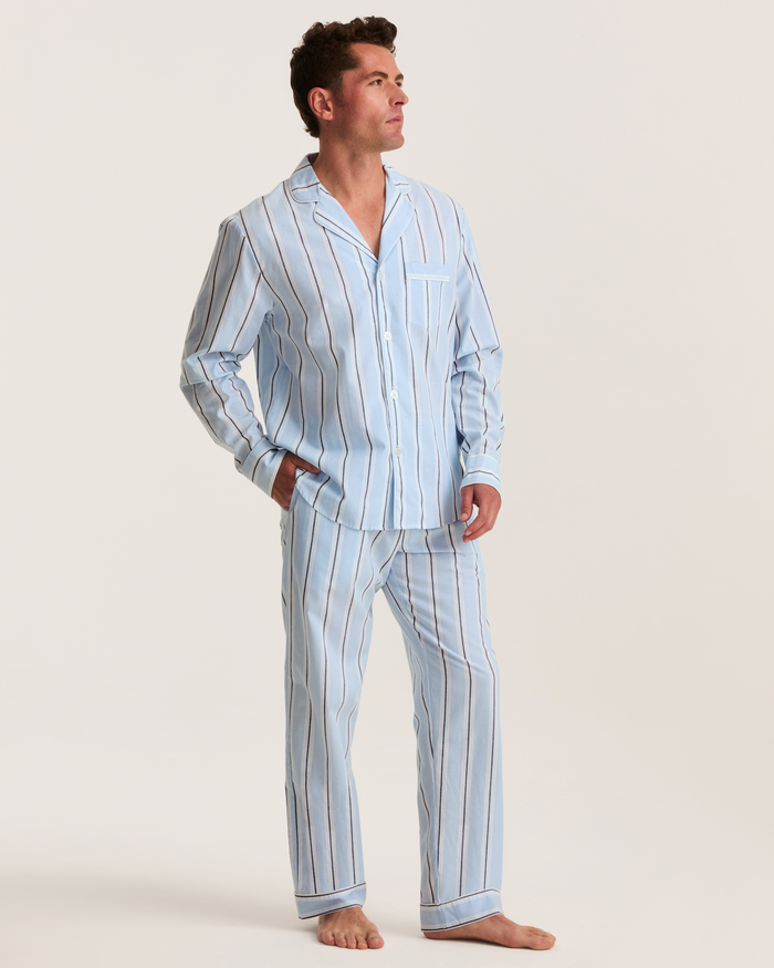 A man stands barefoot, dressed in the Men’s Twill Pajama Set in Coastal Stripe—a light blue set featuring vertical white and dark stripes. With his hands tucked into his pockets, he gazes slightly upward to his left against a plain background.