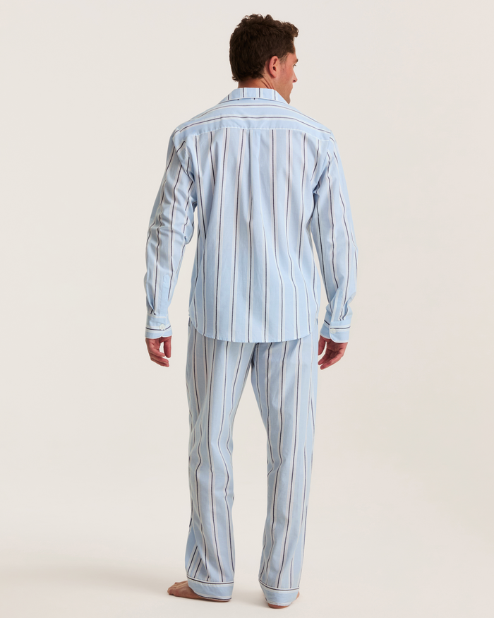 A person stands barefoot facing away, wearing the Men’s Twill Pajama Set in Coastal Stripe—a light blue pajama set with vertical white and dark stripes—against a plain light background.