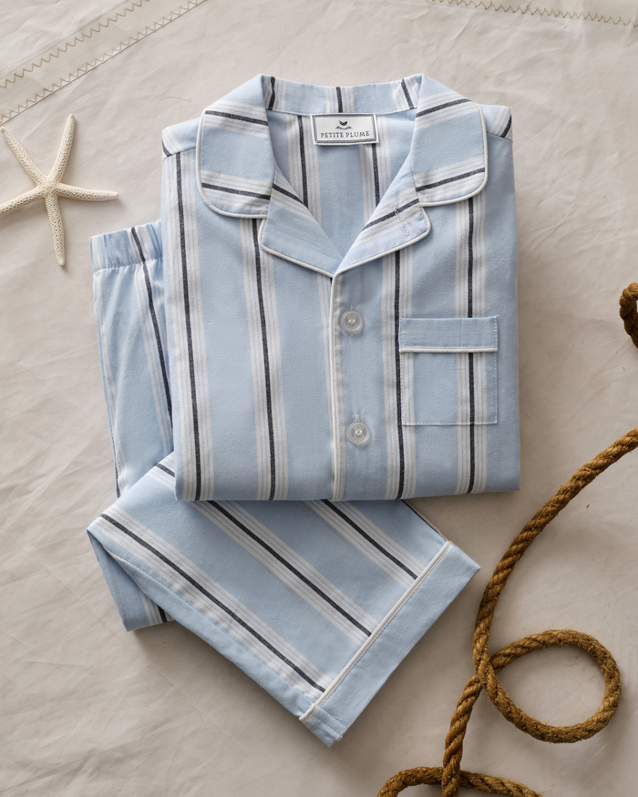 The Men’s Twill Pajama Set in Coastal Stripe, featuring folded light blue stripes and a chest pocket, is displayed on a beige surface. Decorative elements such as a starfish and a piece of thick rope can be seen in the background.