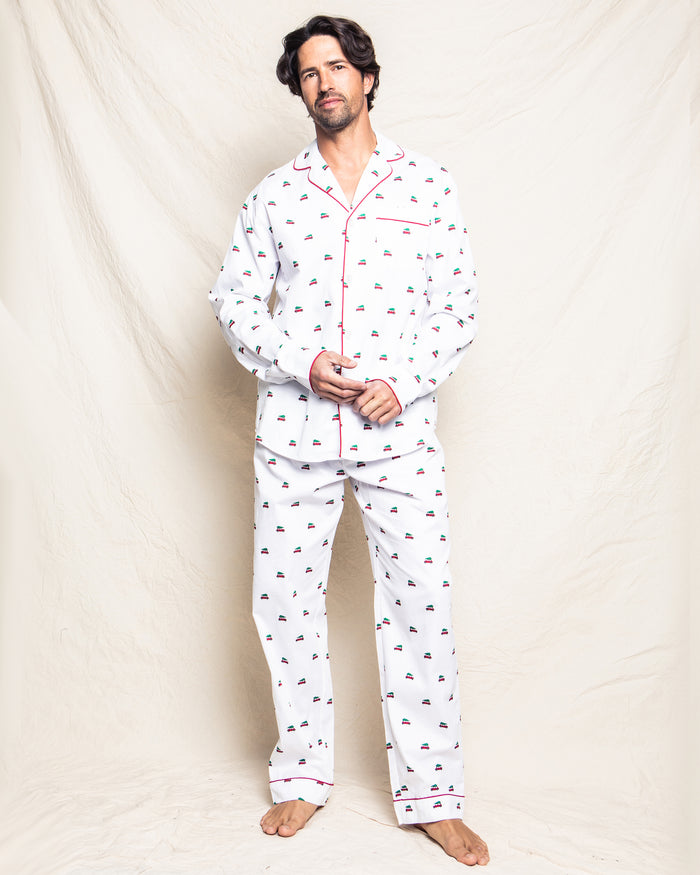 A man with dark hair stands barefoot against a light fabric backdrop, wearing the Petite Plume Mens Twill Pajama Set in Holiday Journey, featuring soft cotton twill with boat prints and red piping for a relaxed look.