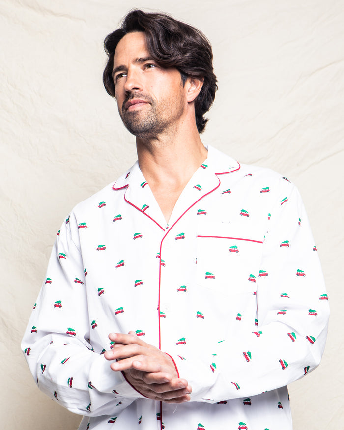 A man with wavy hair wears the Mens Twill Pajama Set in Holiday Journey by Petite Plume. The set features colorful embroidery and red piping on white cotton twill. He stands against a textured light background, hands clasped and gazing sideways, radiating the cozy feel of lazy mornings.