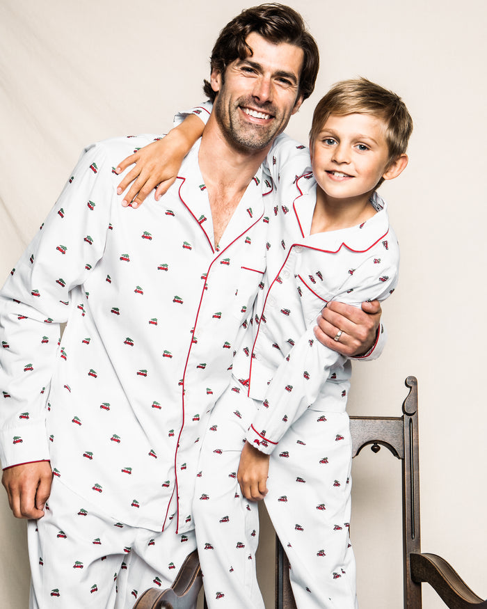 A man and boy are smiling in matching Petite Plume classic pajama sets, featuring small red cars on soft cotton twill. The boy has his arm around the mans shoulder as they stand next to a wooden chair against a neutral background.