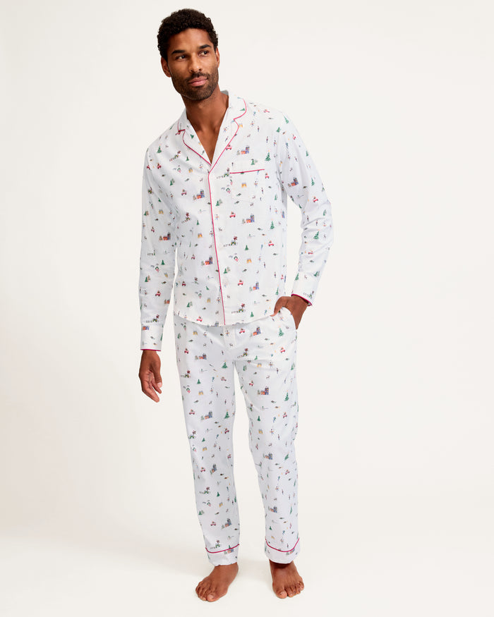 A man stands barefoot against a plain background, wearing the Men's Twill Pajama Set in Holiday Town, featuring a small, colorful print, red piping, a collared button-up top, and long pants.