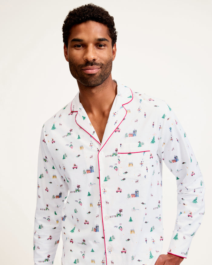 A man stands barefoot against a plain background, wearing the Men's Twill Pajama Set in Holiday Town, featuring a small, colorful print, red piping, a collared button-up top, and long pants.