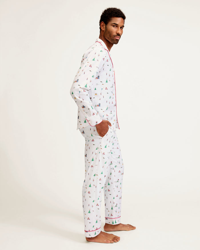 A man stands barefoot against a plain background, wearing the Men's Twill Pajama Set in Holiday Town, featuring a small, colorful print, red piping, a collared button-up top, and long pants.