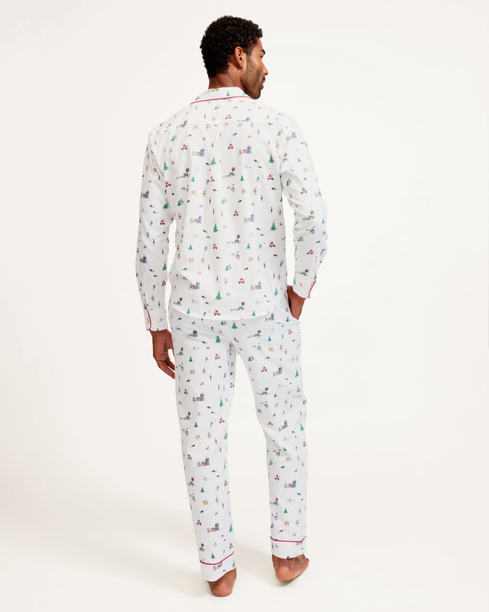 A man stands barefoot against a plain background, wearing the Men's Twill Pajama Set in Holiday Town, featuring a small, colorful print, red piping, a collared button-up top, and long pants.