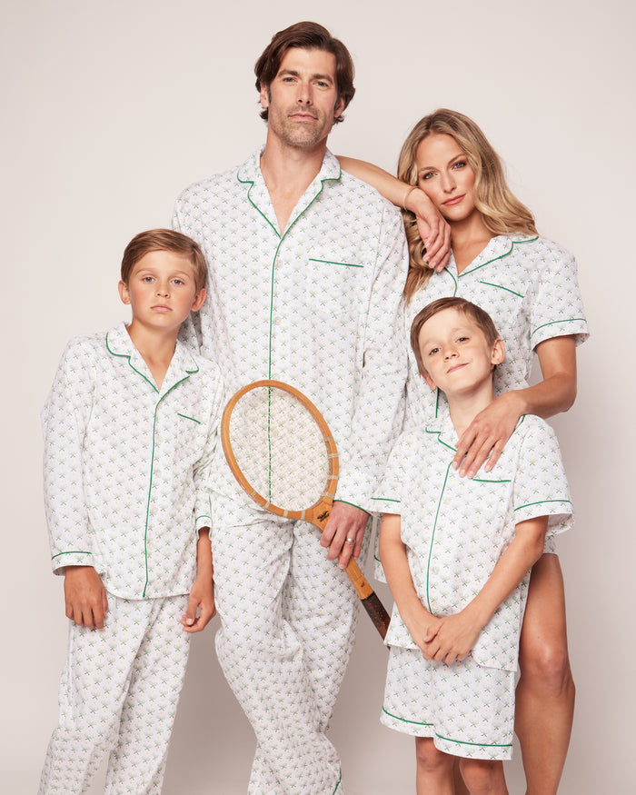 A family of four, wearing Petite Plumes Mens Twill Pajama Set in Match Point, stands together. The father holds a tennis racket beside the mother while two boys pose confidently. Their white pajamas with green piping perfectly blend comfort and style against a plain background.