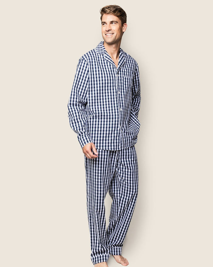 A person stands barefoot in Petite Plumes Mens Twill Pajama Set in Navy Gingham, with short brown hair and a sideward smile, against a plain light beige background.
