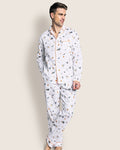 A man models Petite Plumes Mens Twill Pajama Set in Trick or Treat, featuring a playful bird design and orange trim, crafted from premium cotton for comfort and style. He stands against a plain background, hands in pockets and barefoot.