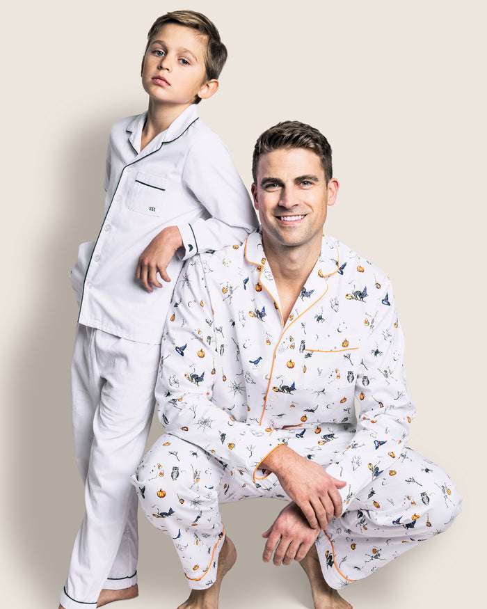 A man and boy in matching Halloween pajamas from Petite Plume pose together; the boy, in white pajamas with black piping, stands with his hand on the mans shoulder, while the man crouches smiling in a Mens Twill Pajama Set featuring a colorful Trick or Treat print.