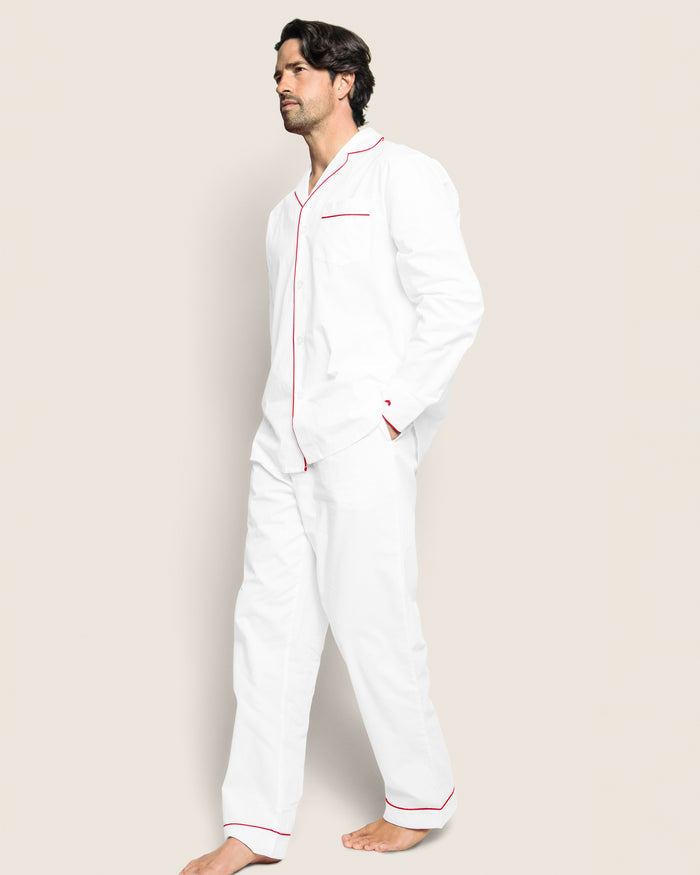 A person stands with hands in pockets, dressed in Petite Plumes Mens Twill Pajama Set in White with Red Piping, crafted from premium cotton. The set includes a collared top and long pants, set against a plain beige background.