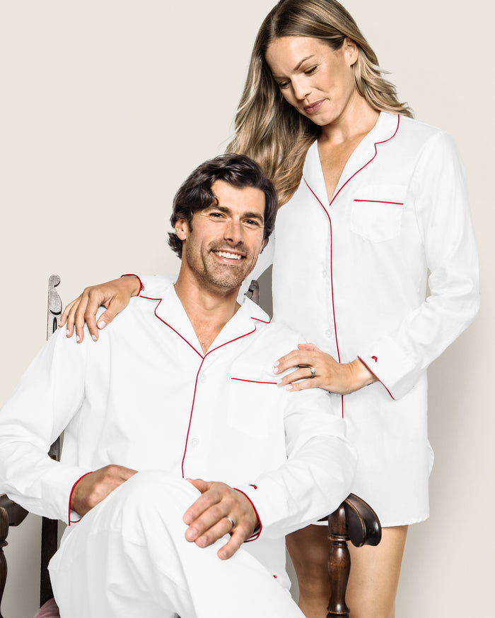 A man sits in a Petite Plume mens twill pajama set—white with red piping—as a woman stands beside him, wearing matching finest-quality cotton sleepwear. He smiles while she rests her hand on his shoulder, looking down slightly.