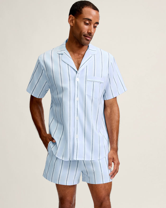 Wearing the Men’s Twill Pajama Short Set in Coastal Stripe—a light blue, short-sleeved ensemble with vertical navy and white stripes, a button-up shirt with chest pocket, and matching shorts—a man stands with one hand in his pocket, looking down.