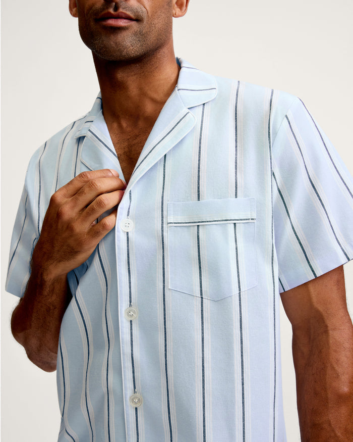 A man wearing the Men’s Twill Pajama Short Set in Coastal Stripe, featuring a light blue, short-sleeve, button-up shirt with dark and white vertical stripes, adjusts his collar.