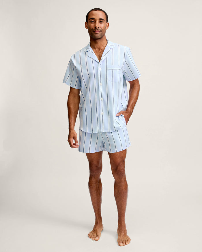 A man stands barefoot, wearing the Men’s Twill Pajama Short Set in Coastal Stripe—a light blue short-sleeved set featuring vertical white and dark blue stripes. The set includes a button-up shirt with a chest pocket and matching shorts.