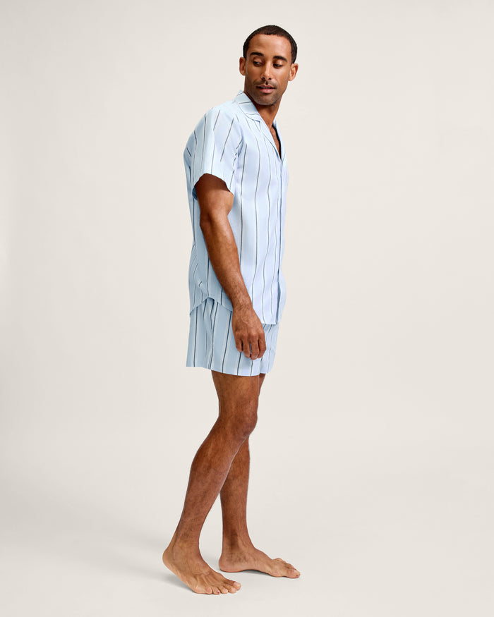 A man stands barefoot on a plain background, wearing the Men’s Twill Pajama Short Set in Coastal Stripe. He looks over his shoulder with a relaxed expression.