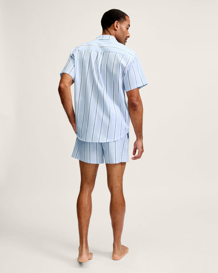 A man stands barefoot facing away, wearing the Men’s Twill Pajama Short Set in Coastal Stripe—a matching light blue short-sleeve shirt and shorts with vertical stripes—against a plain light background.
