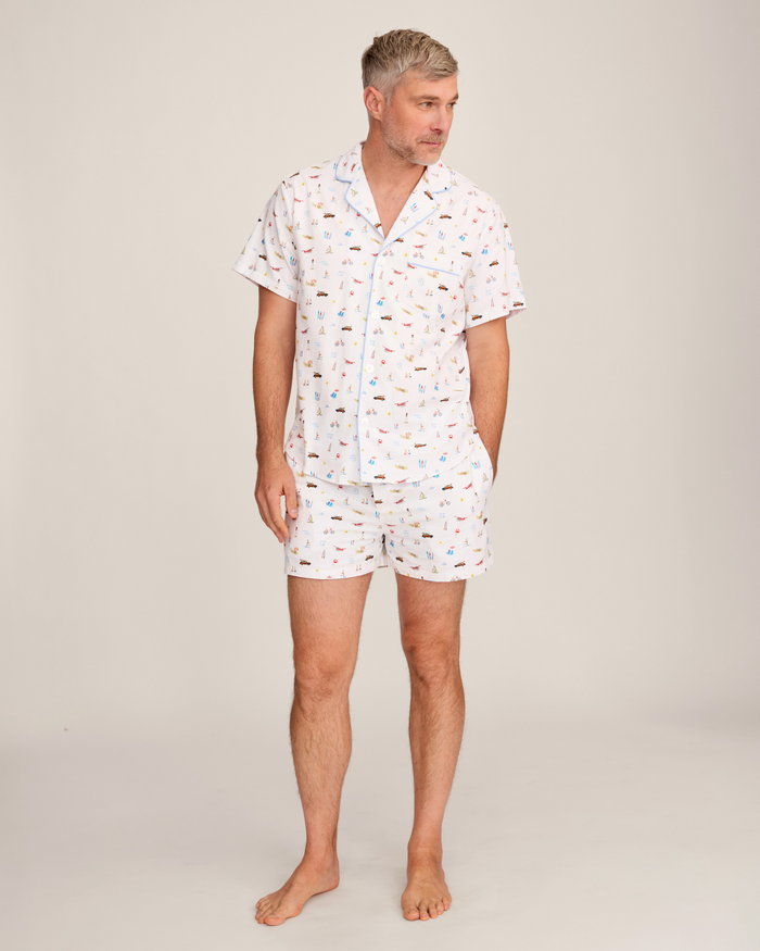 A man stands barefoot, wearing the Men’s Twill Pajama Short Set in Summer Nostalgia—a short-sleeved, button-up shirt and matching shorts with a vibrant print—looking slightly to the side against a plain, light background.
