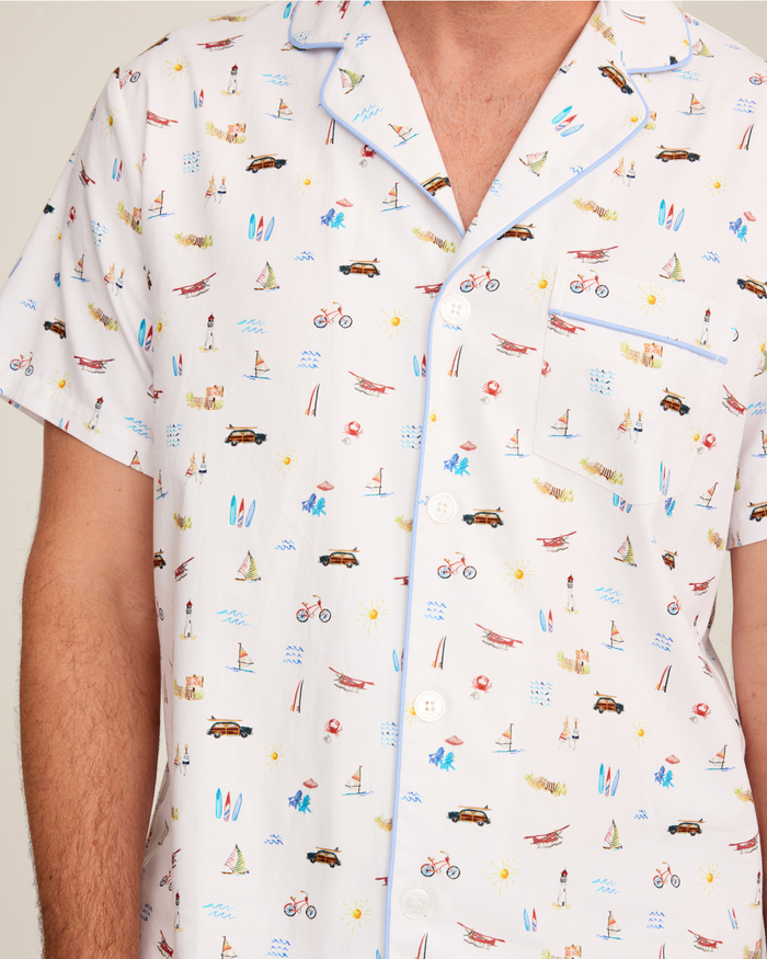 A person is shown from the upper body, wearing the Men’s Twill Pajama Short Set in Summer Nostalgia—a white short-sleeve pajama top featuring small, colorful beach-themed illustrations such as bicycles, cars, surfboards, and lighthouses.