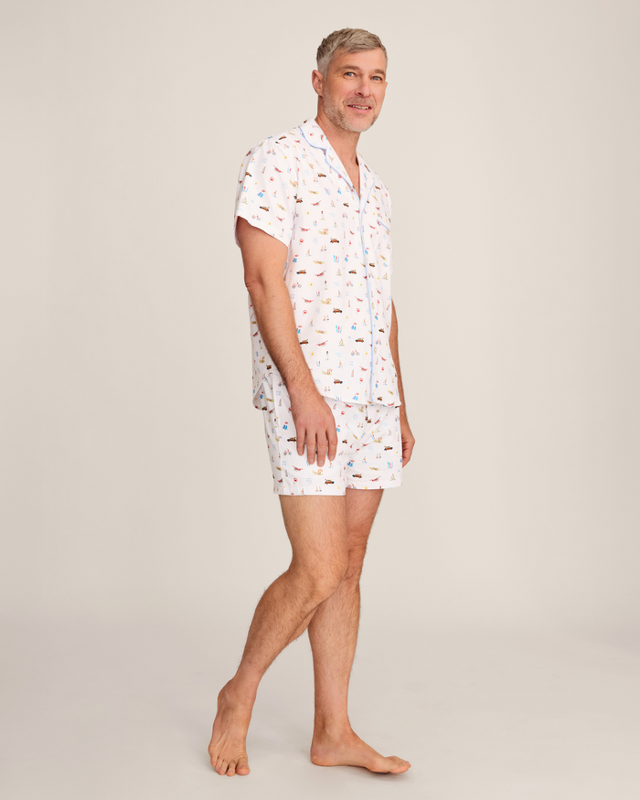 A person with short gray hair wears the Men’s Twill Pajama Short Set in Summer Nostalgia—a matching short-sleeved button-up shirt and shorts with a colorful print—standing barefoot on one leg and smiling against a plain light background.