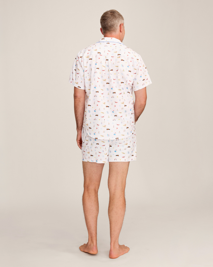 A man stands barefoot facing away, wearing the Men’s Twill Pajama Short Set in Summer Nostalgia—a white short-sleeve shirt and matching shorts with a colorful print—against a plain light background.
