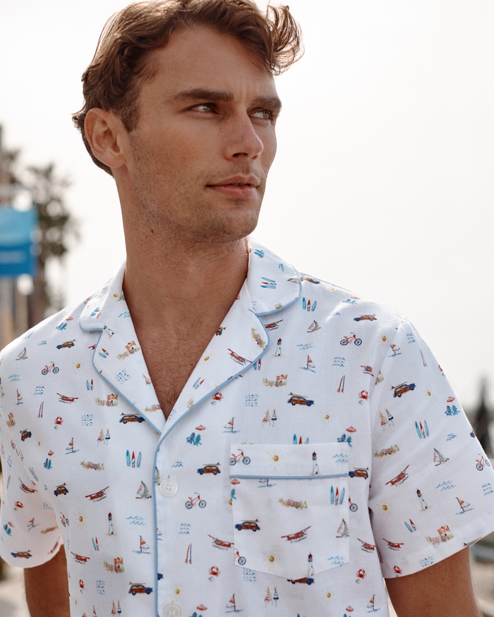 A man stands outdoors, looking to the side, dressed in the Men’s Twill Pajama Short Set in Summer Nostalgia—featuring playful beach and surf illustrations on a white short-sleeve shirt. The bright background appears slightly out of focus.